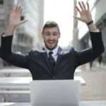 Cheerful businessman raising hands in success gesture in front of laptop, embodying the power of positive wealth affirmations for a prosperous mindset