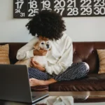 A relaxed individual in cozy home attire cuddles with a serene dog on a leather sofa, with a laptop on a glass table nearby, embodying mindfulness and the joy of simple living.